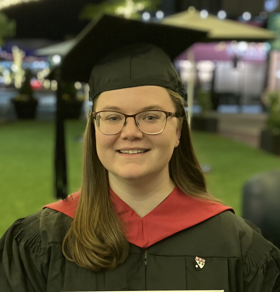 A photo of Kate Daversa. She has shoulder length brown hair, brown metal framed glasses, and green eyes. She is wearing a graduation cap and gown. She is smiling.